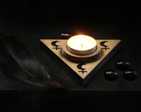 May include: A wooden triangular candle holder with a crescent moon and cross design. A small white candle is burning in the center of the holder. There are black stones on the surface around the holder.