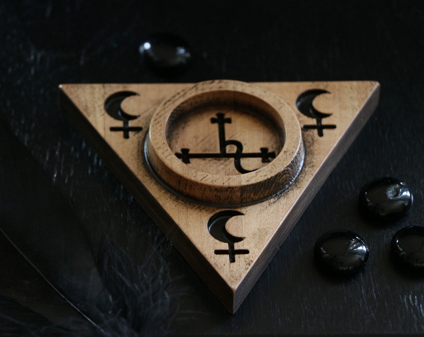 May include: A wooden triangular incense burner with a circular well in the center. The burner is decorated with a pentagram and crescent moon symbols. The burner is sitting on a black surface.