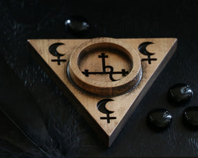 May include: A wooden triangular incense burner with a circular well in the center. The burner is decorated with a pentagram and crescent moon symbols. The burner is sitting on a black surface.