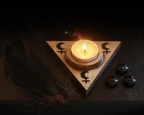 May include: A wooden triangular candle holder with a crescent moon and cross symbols. A small white candle is burning inside the holder. There are black stones and a black feather on a dark surface.