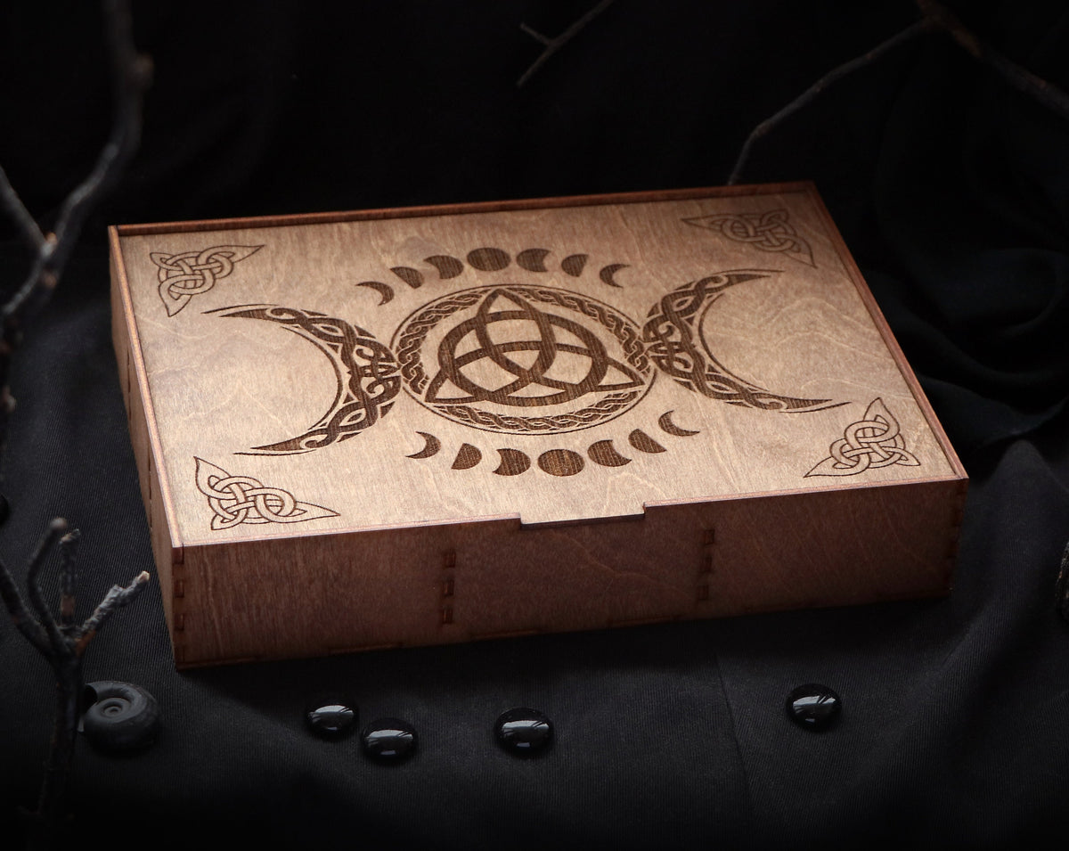 May include: A wooden box with a triple moon and triquetra design. The box is made of light brown wood and has a hinged lid.