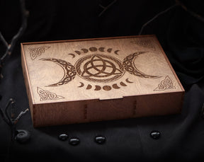 May include: A wooden box with a triple moon and triquetra design. The box is made of light brown wood and has a hinged lid.
