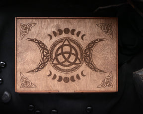 May include: A wooden box with a triple moon and a triquetra design engraved on the lid. The box is brown and has a natural wood finish.