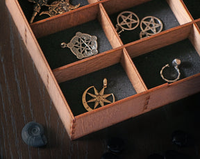 May include: A wooden box with compartments, each containing a different silver or gold pendant. The pendants feature various designs, including stars, circles, and other geometric shapes.