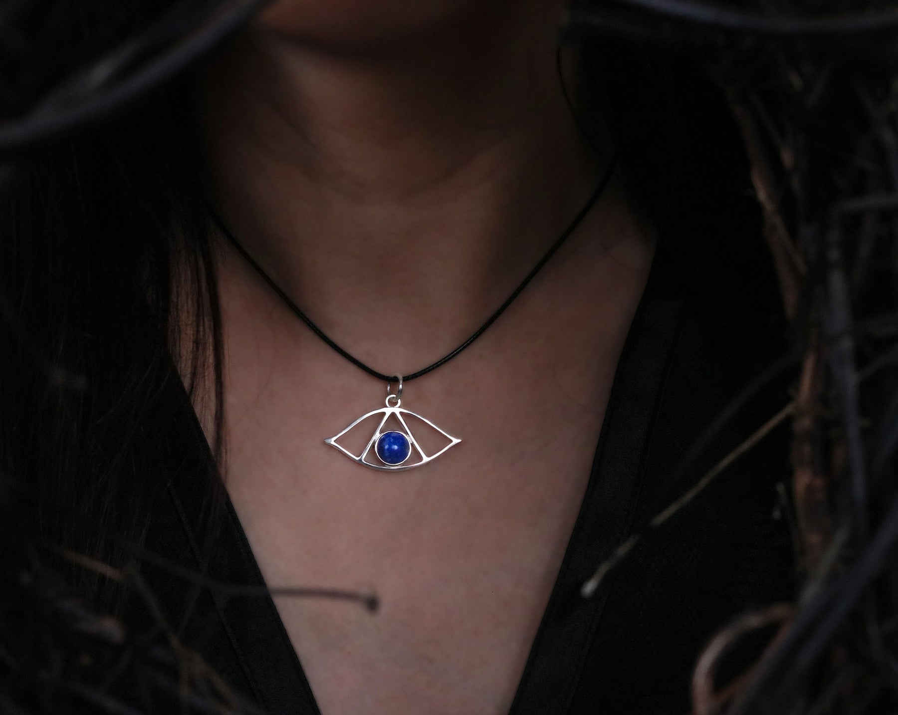 May include: A silver necklace with a geometric pendant featuring a blue gemstone. The pendant is shaped like an eye with a triangular design.