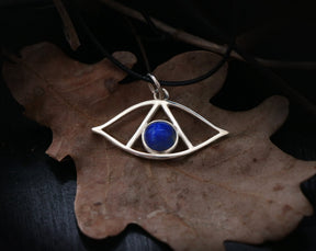 May include: A silver eye pendant with a blue lapis lazuli stone set in the center. The pendant is on a black cord and is resting on a brown leaf.