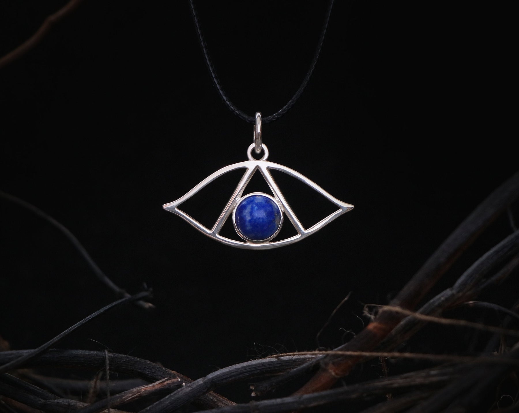 May include: A silver pendant necklace with a blue lapis lazuli stone in the center, shaped like an eye. The pendant is hanging from a black cord.