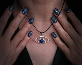 May include: A silver necklace with a blue lapis lazuli stone pendant in the shape of an eye. The necklace is worn by a person with dark blue nail polish.