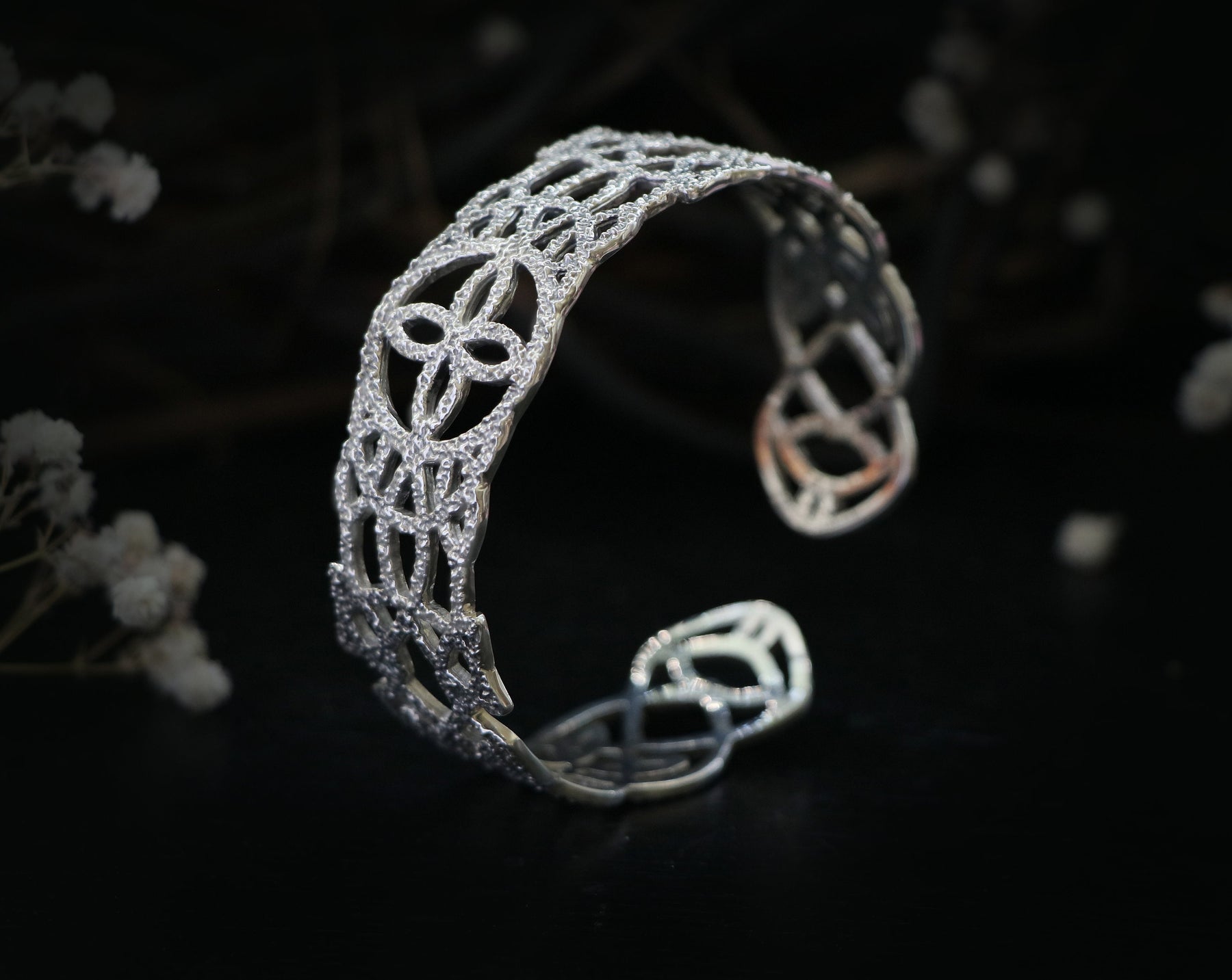 May include: A silver bracelet with a delicate, intricate design. The bracelet is made of a thin metal and has a rounded shape.