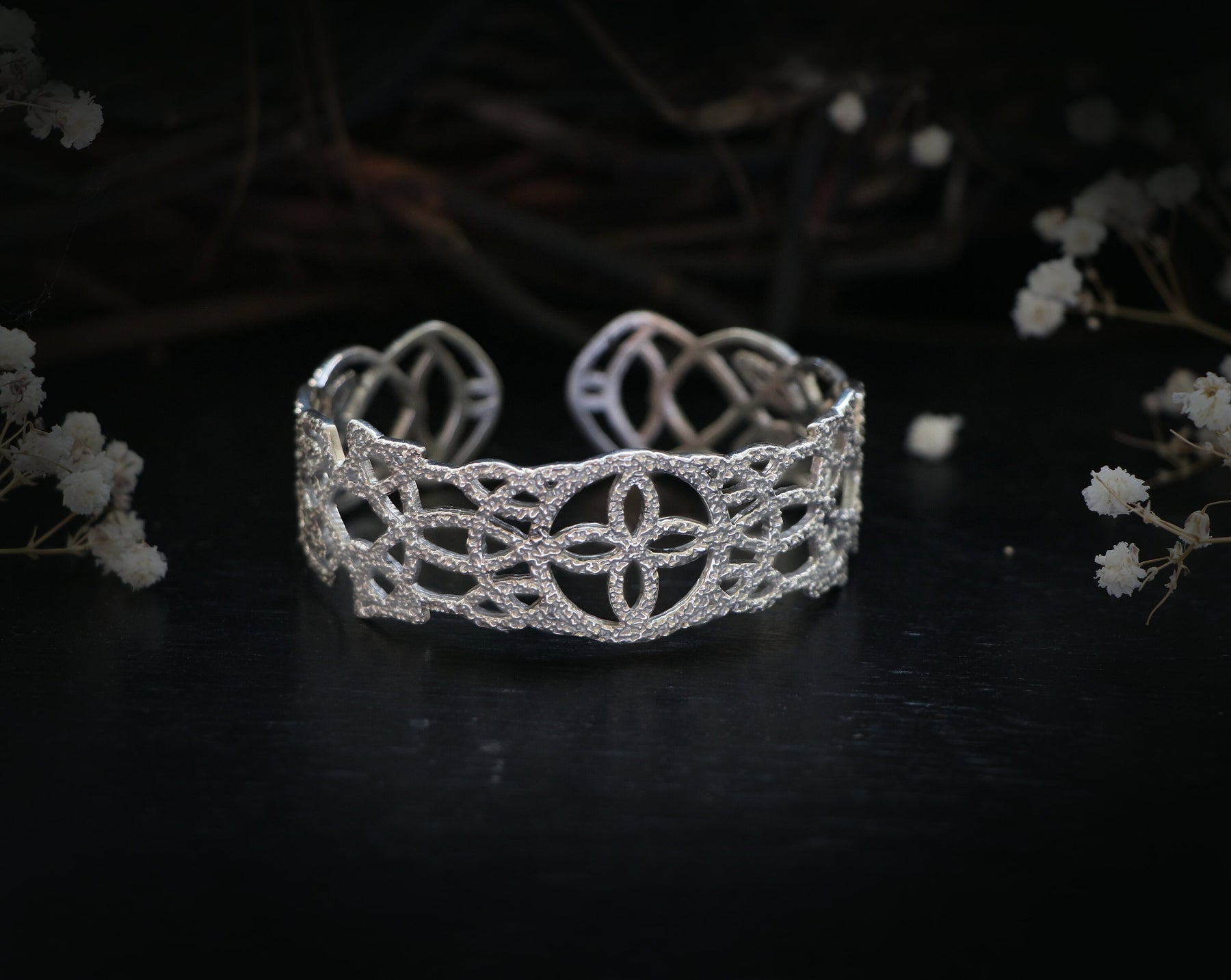 May include: A silver cuff bracelet with a delicate, intricate design. The bracelet is made of a metal with a polished finish and features a repeating pattern of interlocking circles and squares.
