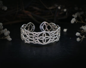 May include: A silver cuff bracelet with a delicate, intricate design. The bracelet is made of a metal with a polished finish and features a repeating pattern of interlocking circles and squares.