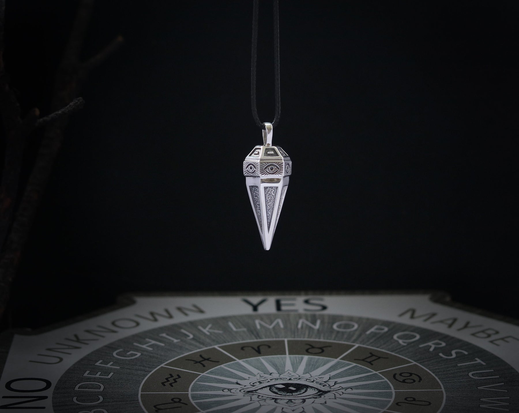 May include: A silver pendulum with a pointed tip and intricate designs hangs from a black cord. The pendulum is positioned above a Ouija board with the letters of the alphabet, numbers, and the words "yes", "no", "maybe", and "unknown".