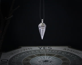 May include: A silver pendulum with a pointed tip and intricate designs hangs from a black cord. The pendulum is positioned above a Ouija board with the letters of the alphabet, numbers, and the words "yes", "no", "maybe", and "unknown".