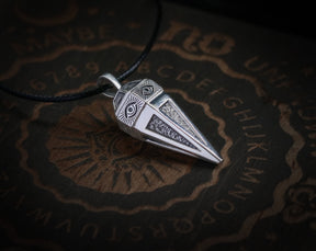 May include: A silver pendant shaped like a pyramid with two eyes on the front. The pendant is hanging from a black cord and is resting on a wooden surface with a Ouija board design.