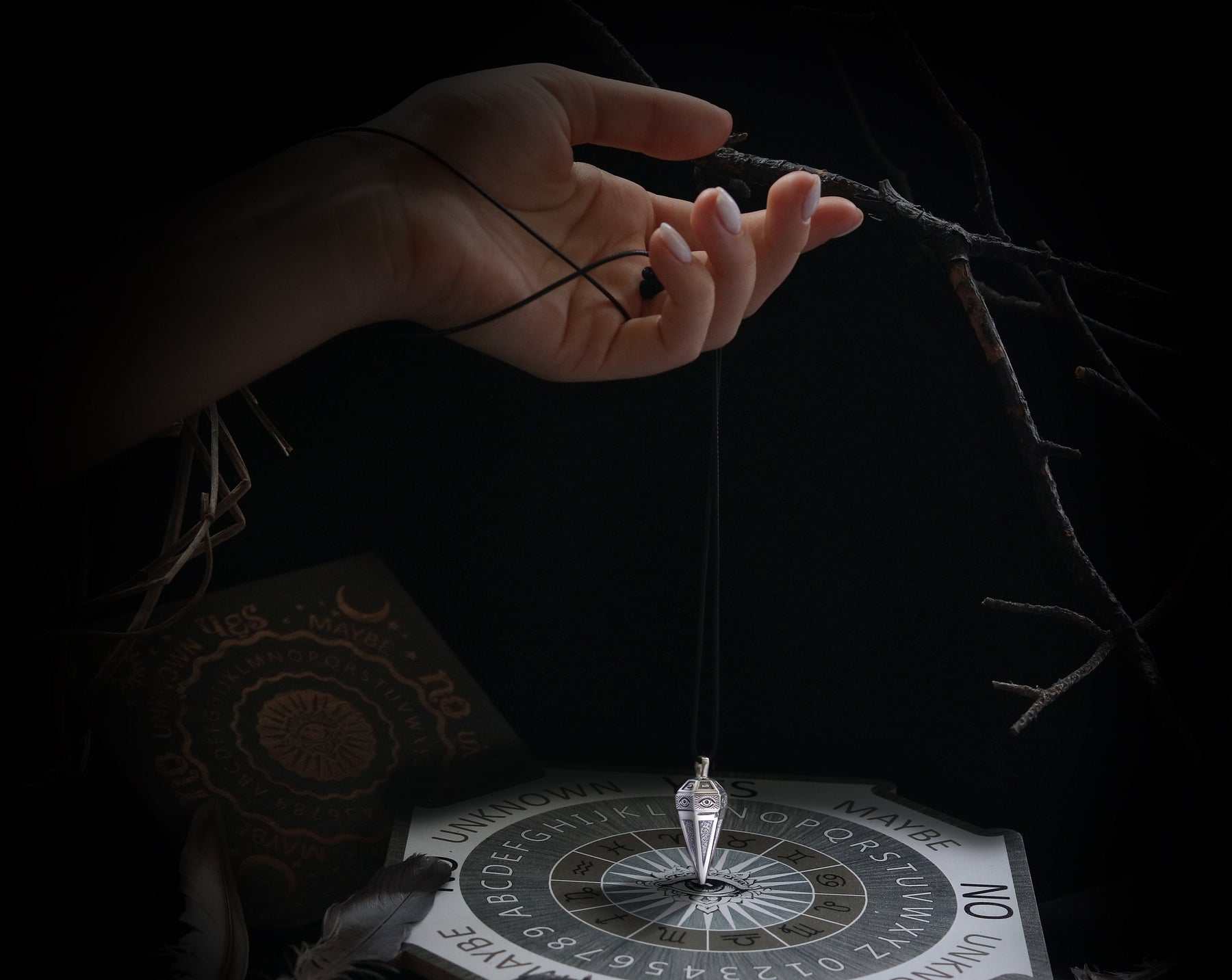 May include: A black and white Ouija board with a silver pendulum hanging above it. The board has letters, numbers, and the words "yes", "no", "maybe", and "unknown".