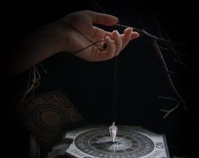 May include: A black and white Ouija board with a silver pendulum hanging above it. The board has letters, numbers, and the words "yes", "no", "maybe", and "unknown".