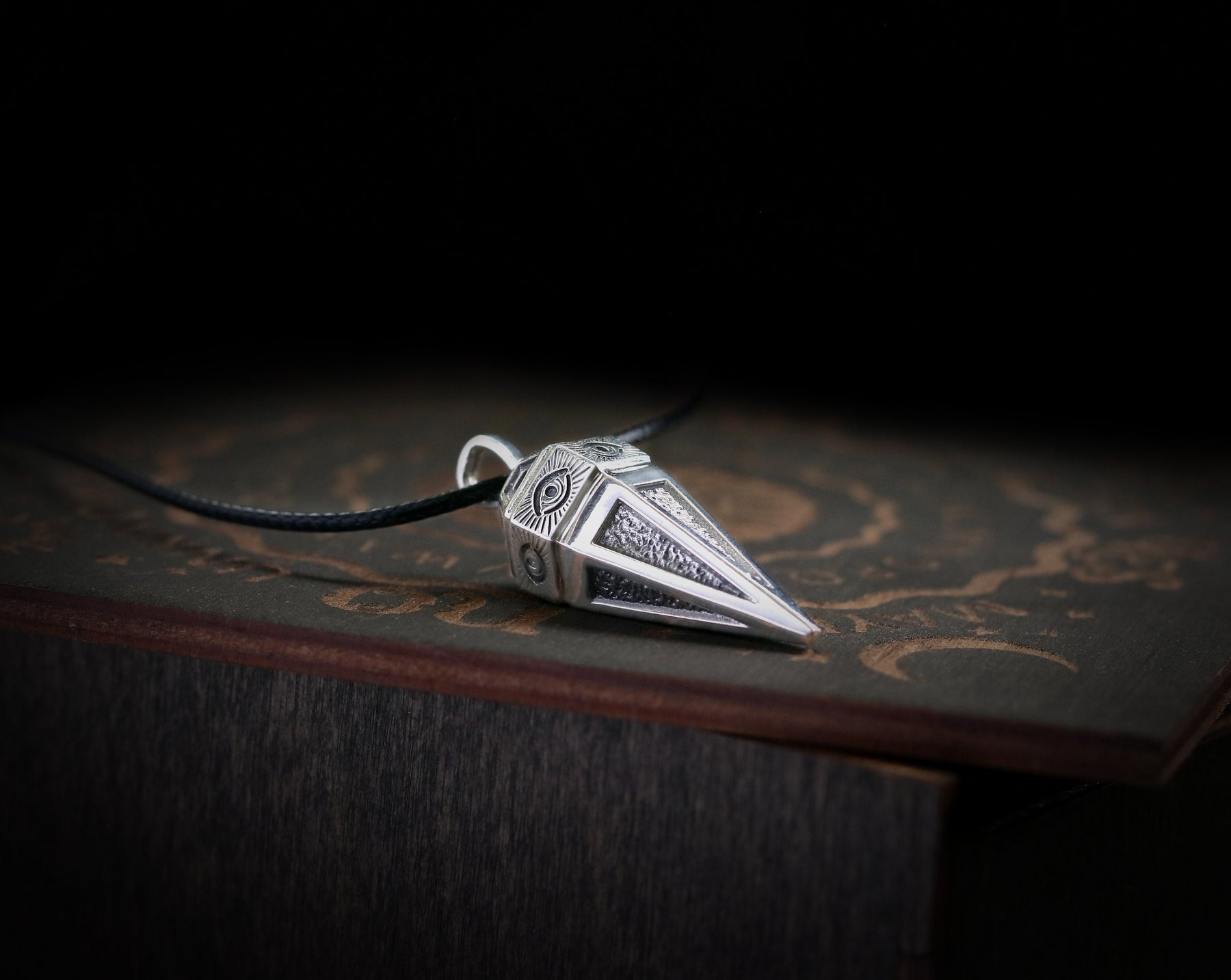 May include: A silver pendant shaped like a pyramid with an eye design on each side. The pendant is on a black cord and is resting on a wooden surface.