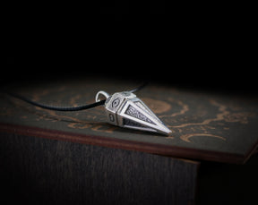 May include: A silver pendant shaped like a pyramid with an eye design on each side. The pendant is on a black cord and is resting on a wooden surface.