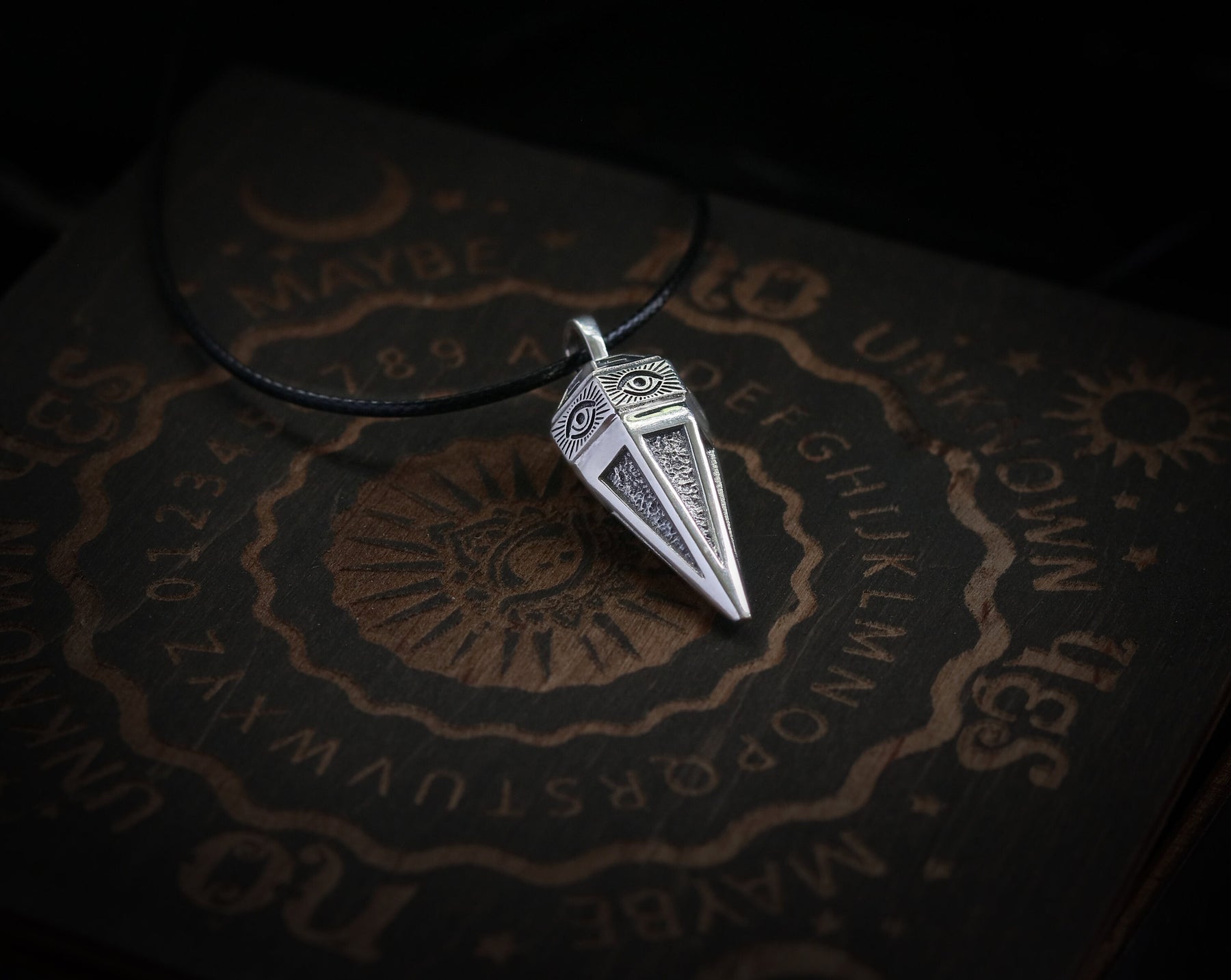 May include: A silver pendant shaped like a pyramid with an eye design on each side. The pendant is on a black cord and is resting on a wooden surface with a circular design and the letters of the alphabet.