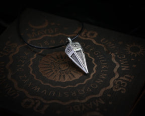 May include: A silver pendant shaped like a pyramid with an eye design on each side. The pendant is on a black cord and is resting on a wooden surface with a circular design and the letters of the alphabet.
