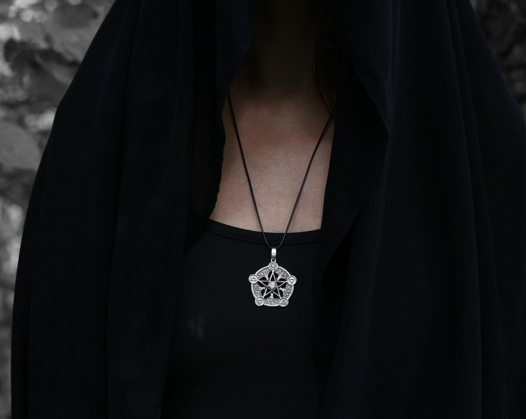 May include: A silver pendant necklace with a detailed star design. The necklace is worn by a person wearing a black hooded garment.