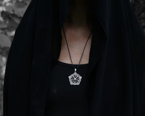 May include: A silver pendant necklace with a detailed star design. The necklace is worn by a person wearing a black hooded garment.
