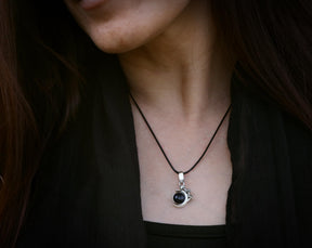May include: A black gemstone pendant necklace with a silver setting. The necklace is on a black cord and is worn by a person.