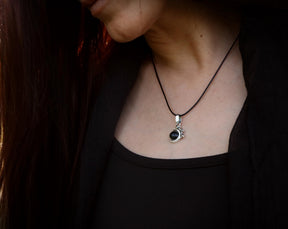 May include: A silver necklace with a black gemstone pendant. The pendant is a round, cabochon-cut stone set in a silver setting. The necklace is on a black cord.