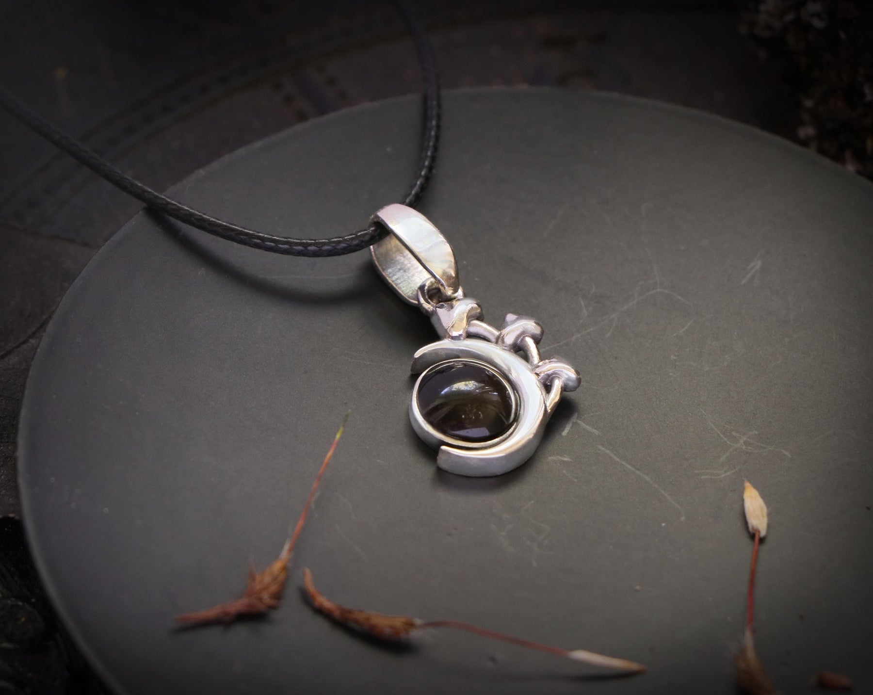 May include: A silver pendant necklace with a dark gray gemstone set in a crescent moon design. The pendant is on a black cord.