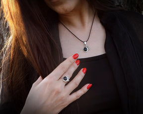 May include: A woman wearing a black top with a silver necklace and pendant featuring a black stone. She is also wearing a silver ring with a black stone on her finger.