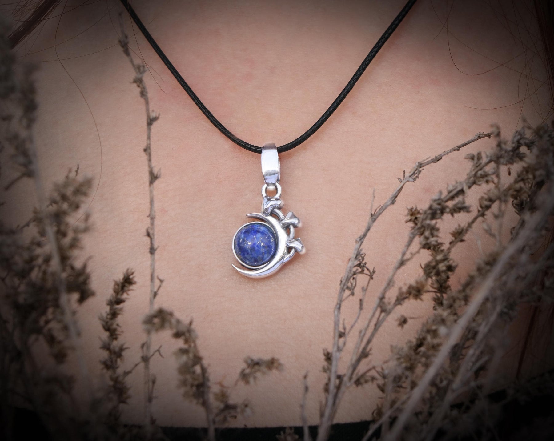 May include: A silver crescent moon pendant necklace with a blue lapis lazuli stone set in the center. The necklace is on a black cord.