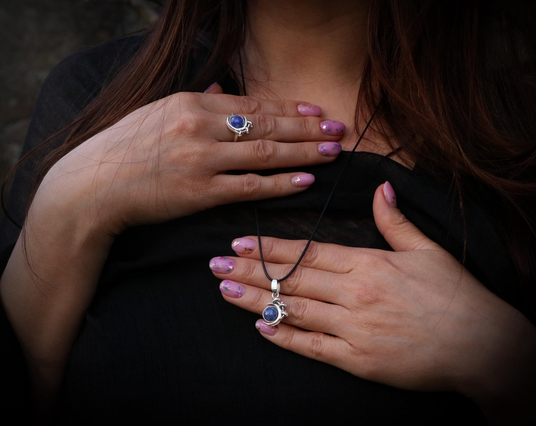 May include: A silver necklace and ring set with a blue gemstone pendant and ring. The necklace has a black cord and the ring is on a finger with pink nail polish.