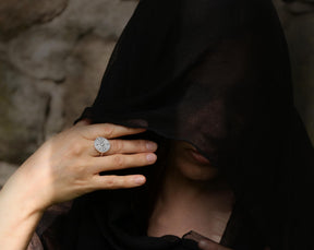 A mysterious lifestyle photograph of a woman shrouded in a black fabric veil that covers her eyes. Her hand is raised to her face, prominently displaying the sterling silver Wheel of Hecate (Strophalos) ring. The silver and central gemstone contrast sharply against the dark veil, creating a powerful occult and witchy aesthetic. Crafted by Pagan Workshop.